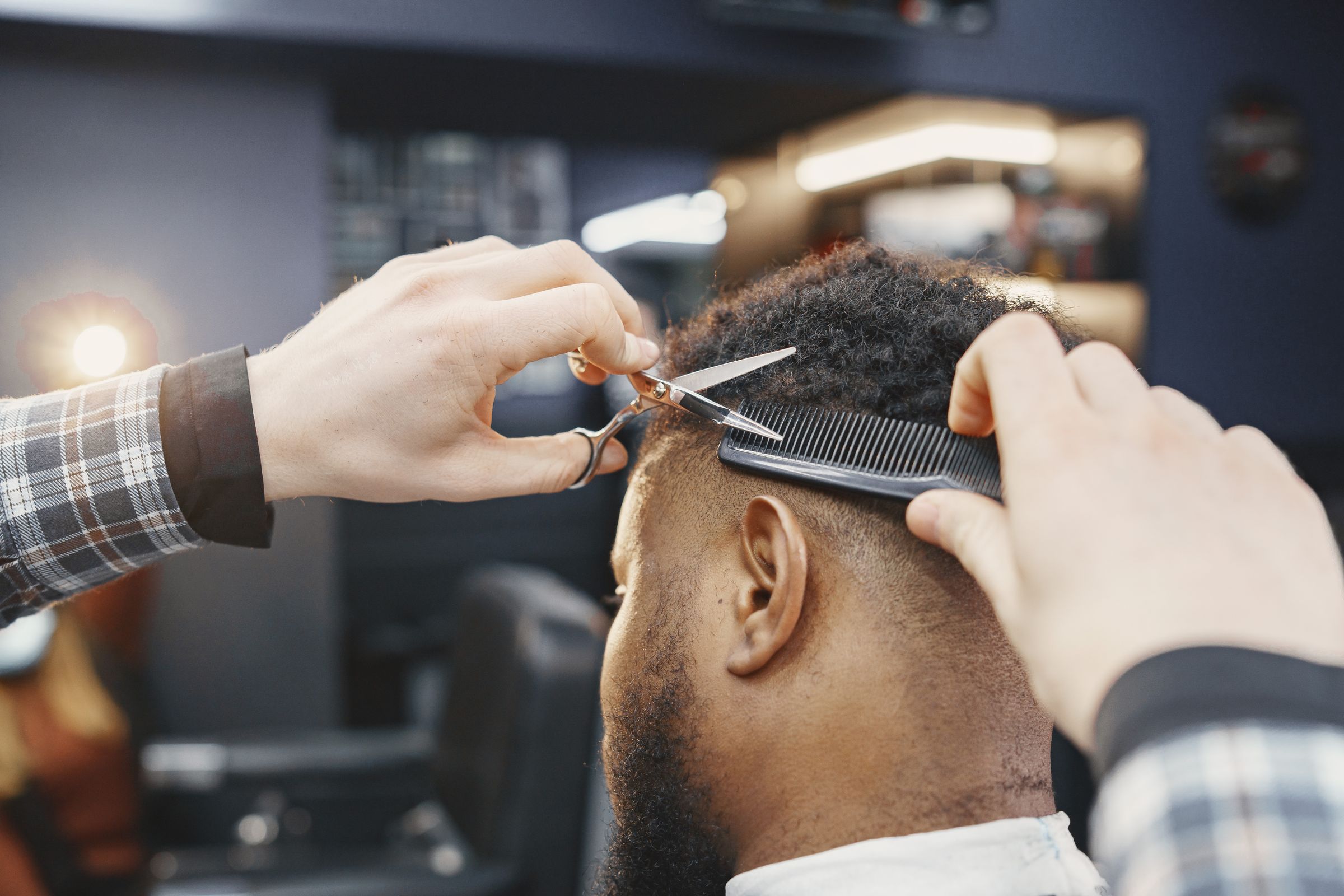 A barber's hands cut a Black man's short curly hair with scissors and a comb, seen from behind in a softly lit barbershop.